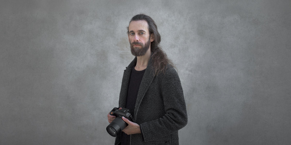 Long-haired bearded photographer holding a Hasselblad camera with a large lens against a textured gray concrete wall