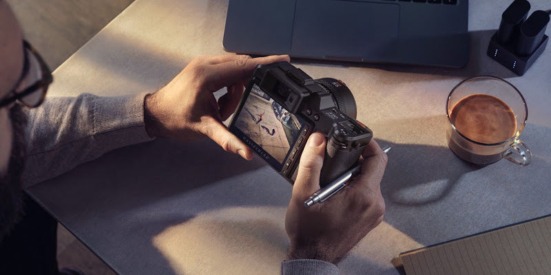 A photographer reviewing an image on a Hasselblad medium format camera beside a laptop, coffee cup, and notebook on a desk
