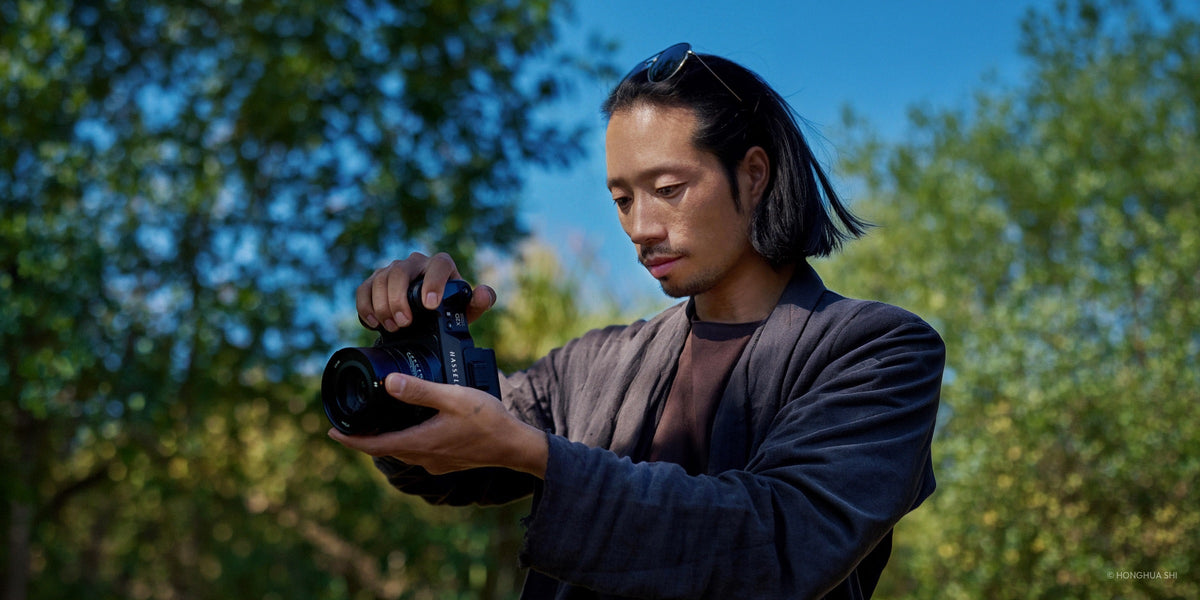 Photographer adjusting a Hasselblad X2D camera outdoors against a backdrop of sunlit trees